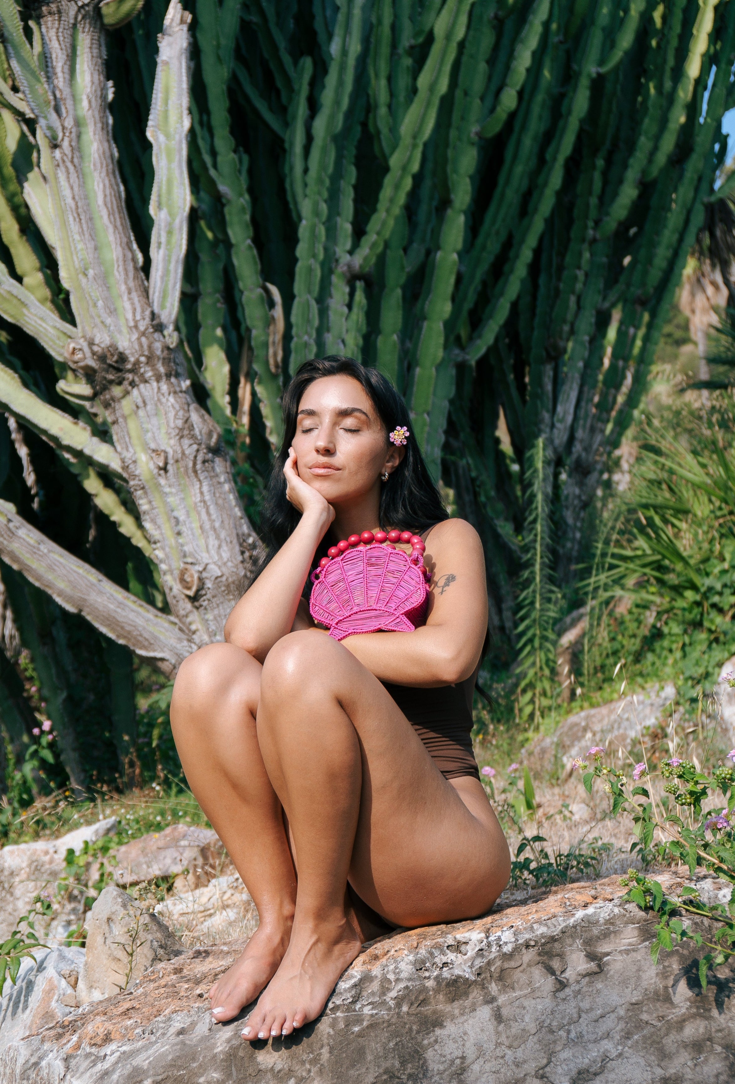 Woman sitting on a rock with a cactus plant in the background and a shell hanbag