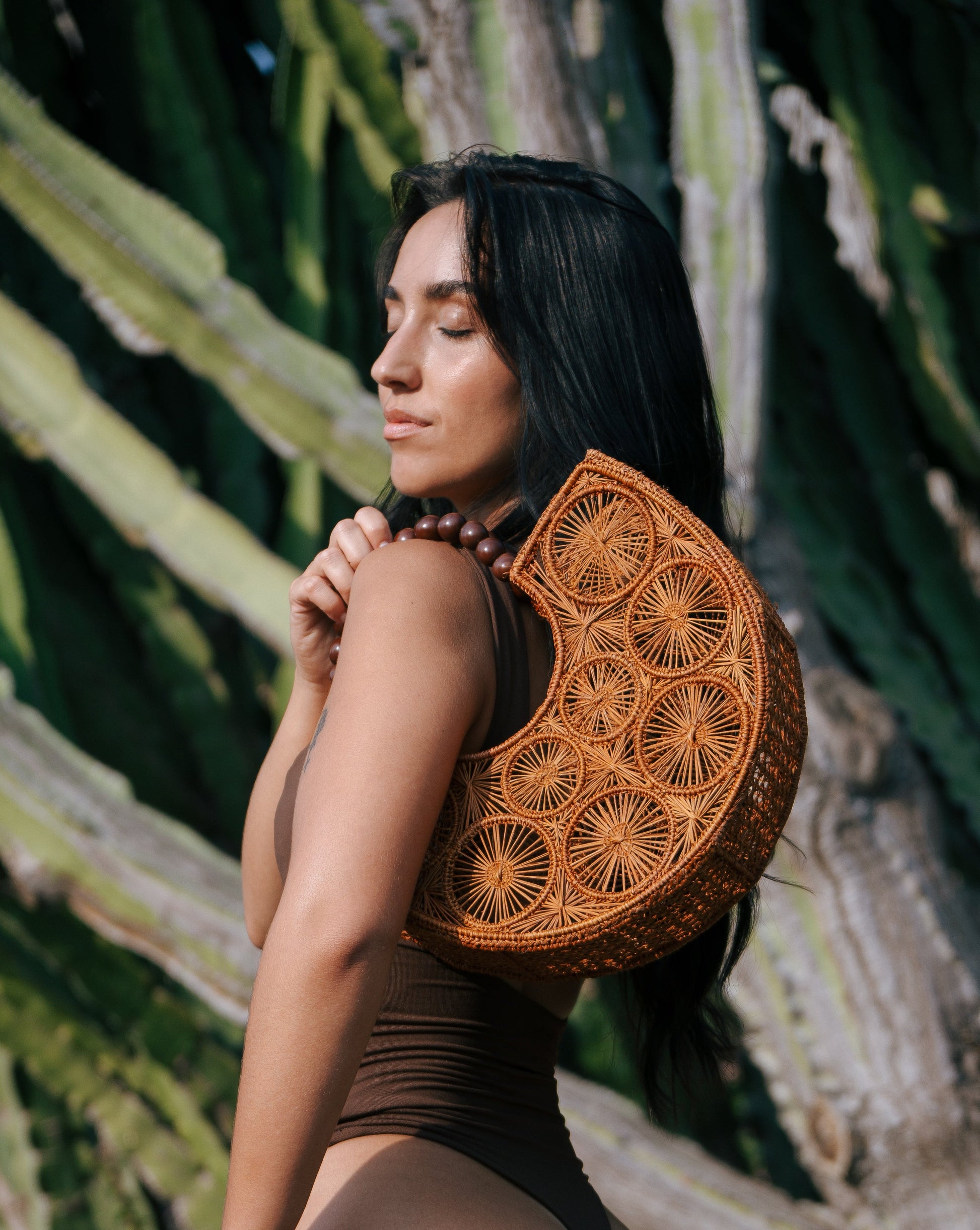 Woman wearing a brown swimsuit with a decorative woven bag Maison Lana piece against a natural background